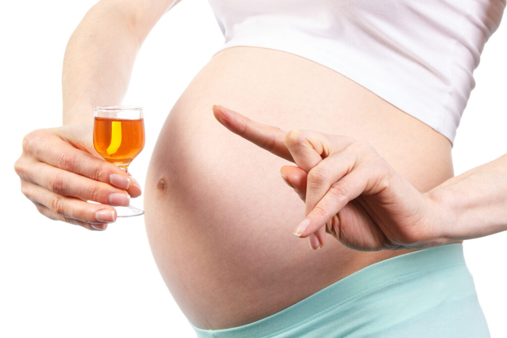 Pregnant woman gesturing towards her belly and a glass of alcohol, illustrating addiction risk factors.