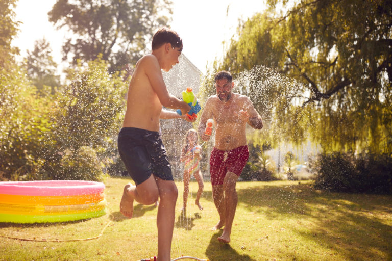 Family Wearing Swimming Costumes Having Water Fight With Water Pistols In Summer Garden